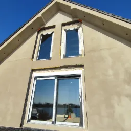 Montaż okien w nowym domu z oknem przesuwnym na parterze, widoczne pianki montażowe i folie ochronne na ramach, elewacja w trakcie wykończenia, błękitne niebo w tle.