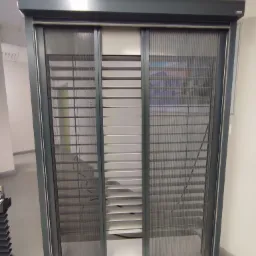 Szara przesuwna moskitiera plisowana, widok z bliska na prowadnice i siatkę, z widocznymi lamelami żaluzji w tle.