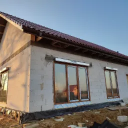 Dom w trakcie budowy z zamontowanymi oknami z roletami zewnętrznymi, widoczne bloczki betonowe i częściowo ułożona dachówka.