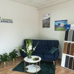 IJM OKNA - Salon firmowy z ekspozycją bram garażowych Nice na stojaku, ciemnoniebieska sofa, kwiaty w wazonie na stoliku, logo firmy IJM Łuczkowiec Czarnecki na ścianie, plakaty reklamowe rolet i napędów Nice.