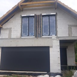 IJM OKNA - Surowy stan budynku jednorodzinnego w trakcie budowy, z zamontowaną czarną bramą garażową segmentową i dwoma oknami z białymi ramami na piętrze, widoczne drewniane elementy konstrukcji dachu.