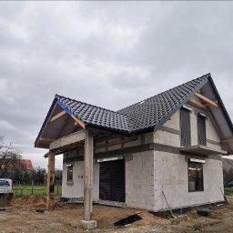 IJM OKNA - Surowy stan domu jednorodzinnego z czarną dachówką i zamontowanymi roletami zewnętrznymi, widok z zewnątrz na plac budowy z samochodem dostawczym w tle.