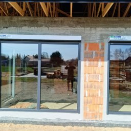 IJM OKNA - Szare okna aluminiowe z roletami zewnętrznymi w surowym stanie budynku. Widoczne cegły i drewniana konstrukcja dachu. W oknach odbija się krajobraz i osoba robiąca zdjęcie.