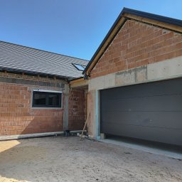 IJM OKNA - Szara brama garażowa segmentowa w surowym otworze budynku z cegły, z widocznym oknem i dachem. Budowa w toku w słoneczny dzień.