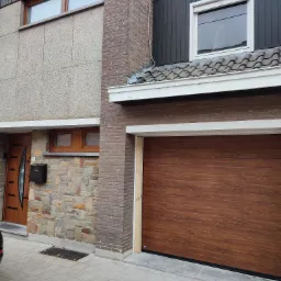 Brązowa segmentowa brama garażowa z efektem drewna, zamontowana w nowoczesnym domu z cegły i kamienia, widok z zewnątrz na podjazd.