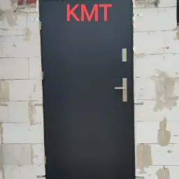 Ciemnoszare drzwi z klamką i zabezpieczeniem, osadzone w surowej ścianie z białych bloczków, uszczelnione pianką montażową i z czerwonym napisem KMT u góry.