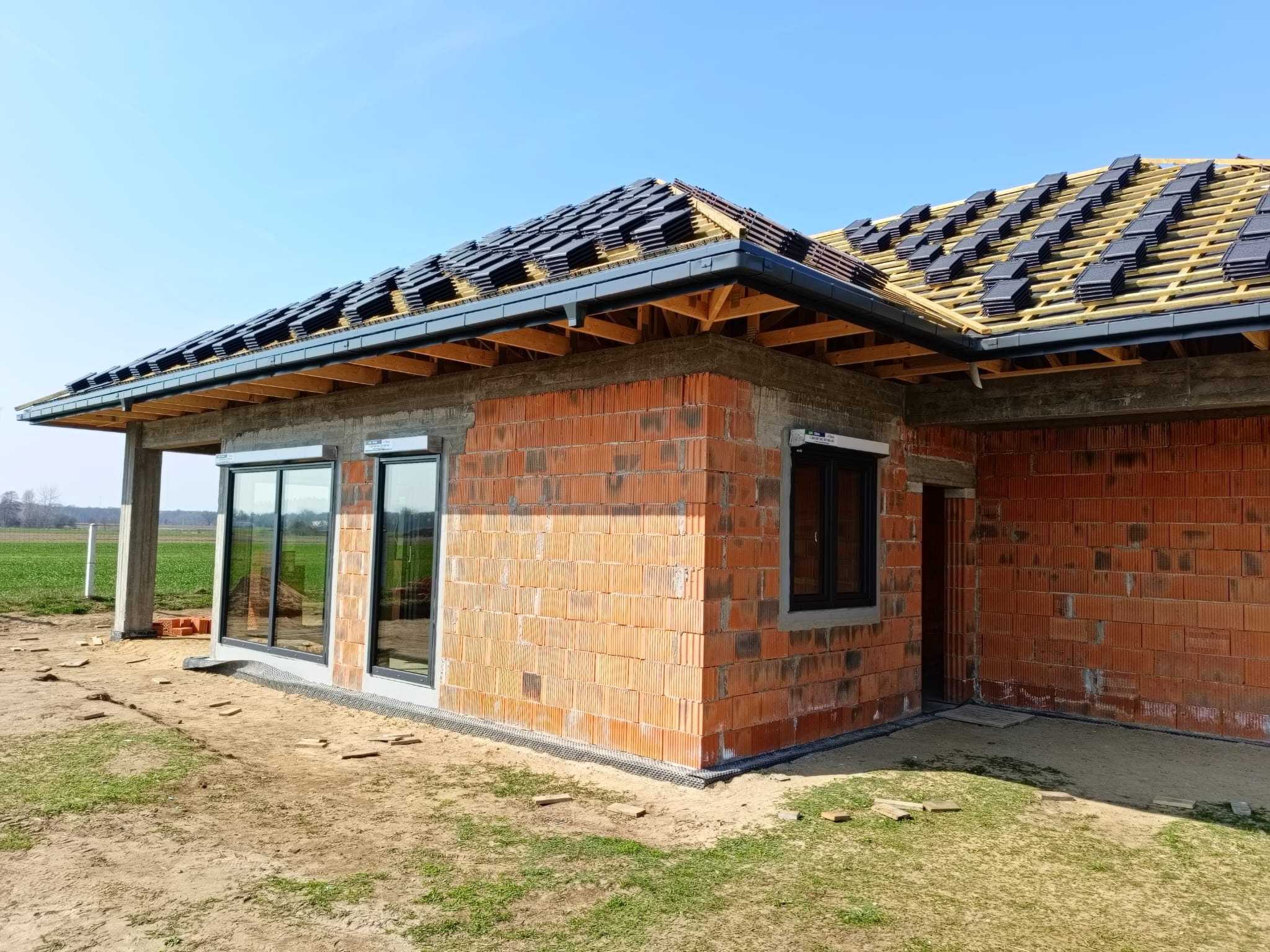 Budowa domu jednorodzinnego: ściany z czerwonej cegły, czarne okna, dach w trakcie krycia dachówką na tle błękitnego nieba i zielonego pola.