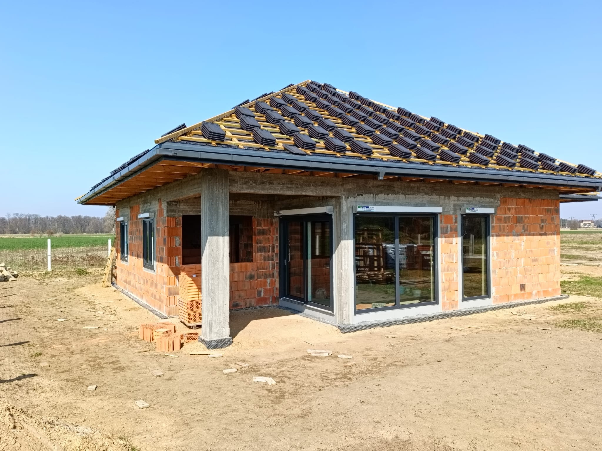 Budowa domu jednorodzinnego w stanie surowym otwartym: mury z cegły, okna z roletami zewnętrznymi, dach z dachówką w trakcie układania, błękitne niebo.