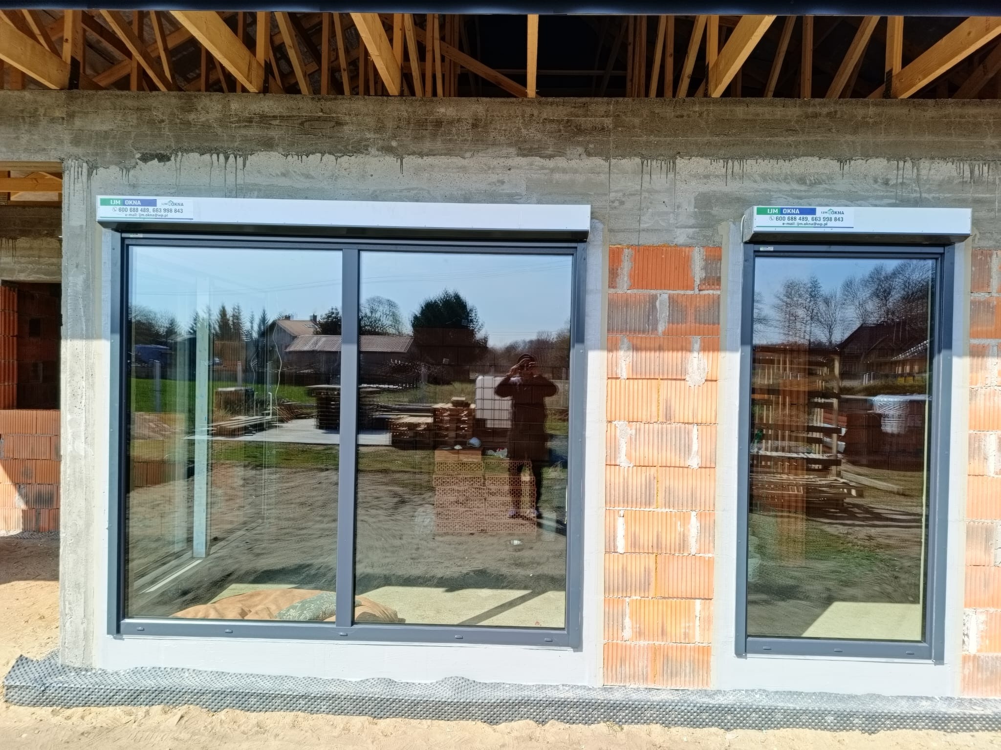 Szare okna aluminiowe z roletami zewnętrznymi w surowym stanie budynku. Widoczne cegły i drewniana konstrukcja dachu. W oknach odbija się krajobraz i osoba robiąca zdjęcie.