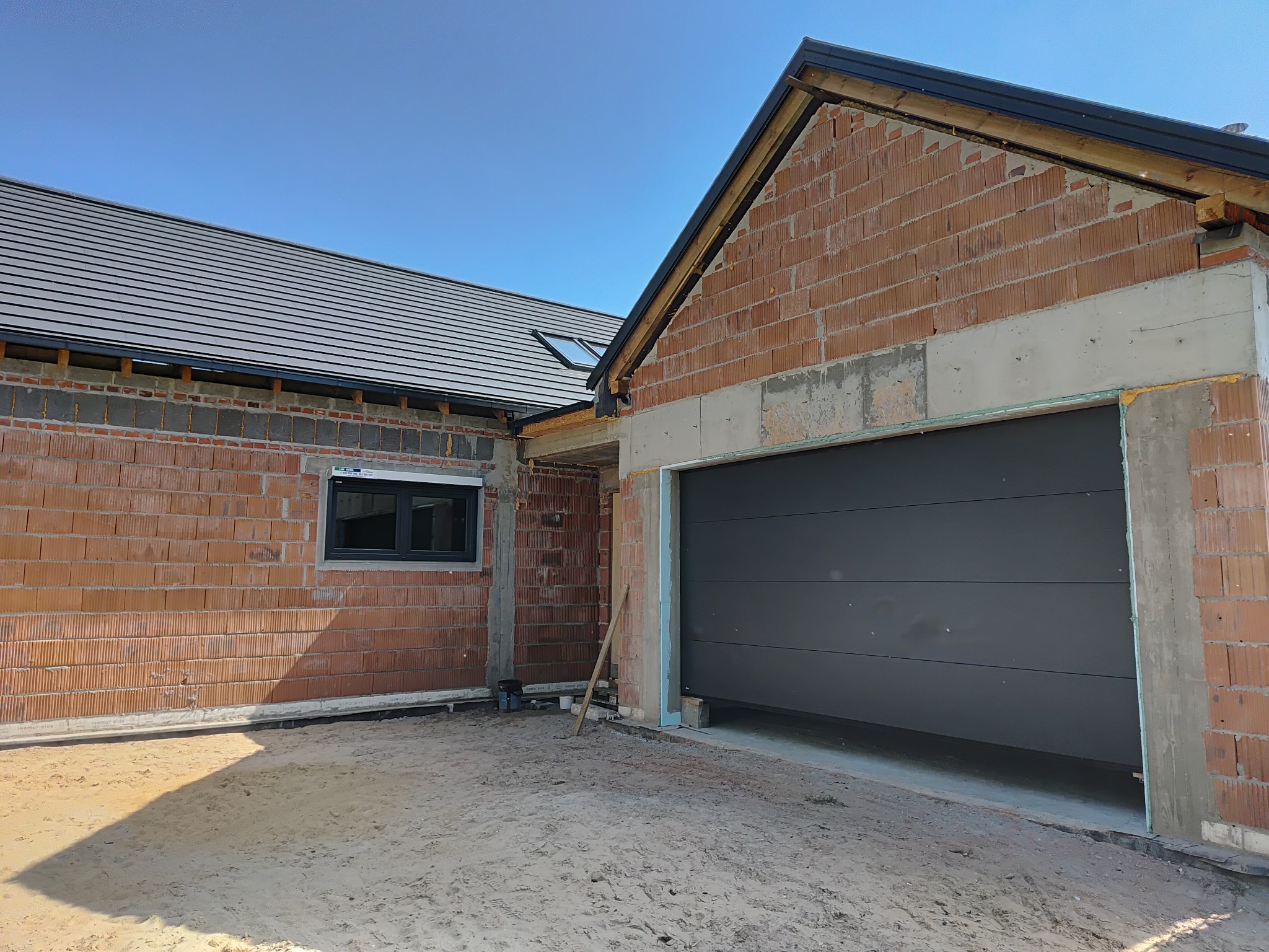 Szara brama garażowa segmentowa w surowym otworze budynku z cegły, z widocznym oknem i dachem. Budowa w toku w słoneczny dzień.