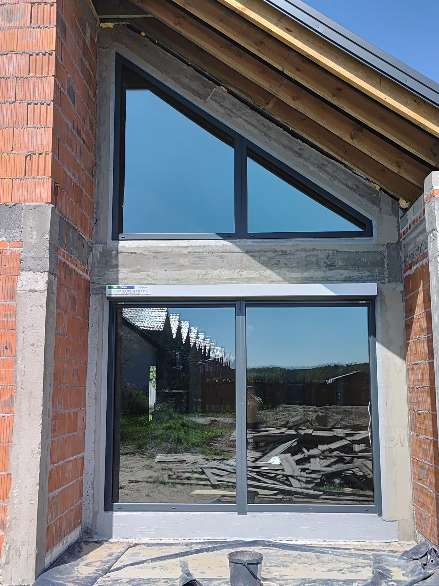 Aluminiowe okno z roletą zewnętrzną w surowym stanie budynku z cegły. Nietypowy kształt okna trójkątnego. Widoczne odbicie otoczenia w szybie.