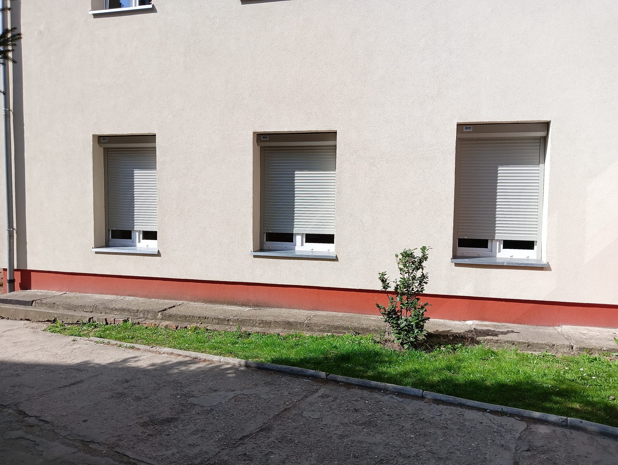 Elewacja budynku z trzema oknami zabezpieczonymi roletami zewnętrznymi w kolorze szarym. Widoczne fragmenty okien i parapety. Na dole trawnik i chodnik.