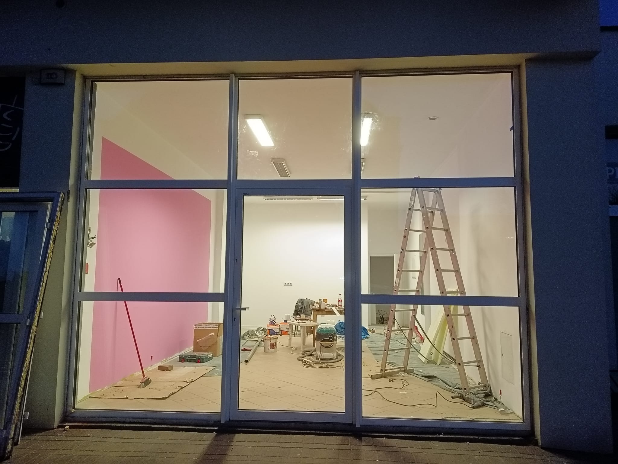 Duże okno aluminiowe z widokiem na wnętrze w trakcie remontu. Widoczna drabina, narzędzia malarskie i fragment pomalowanej na różowo ściany.