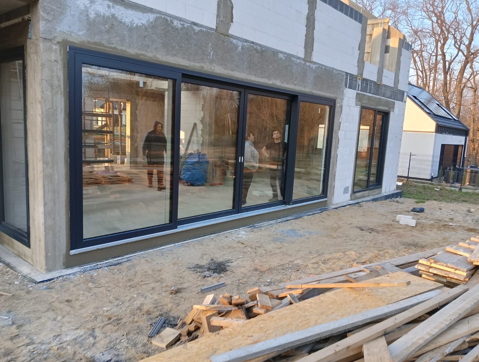 Nowoczesne, przesuwne okna aluminiowe w surowym budynku z betonu komórkowego. Widoczne refleksy i osoby wewnątrz, drewniane deski na zewnątrz.