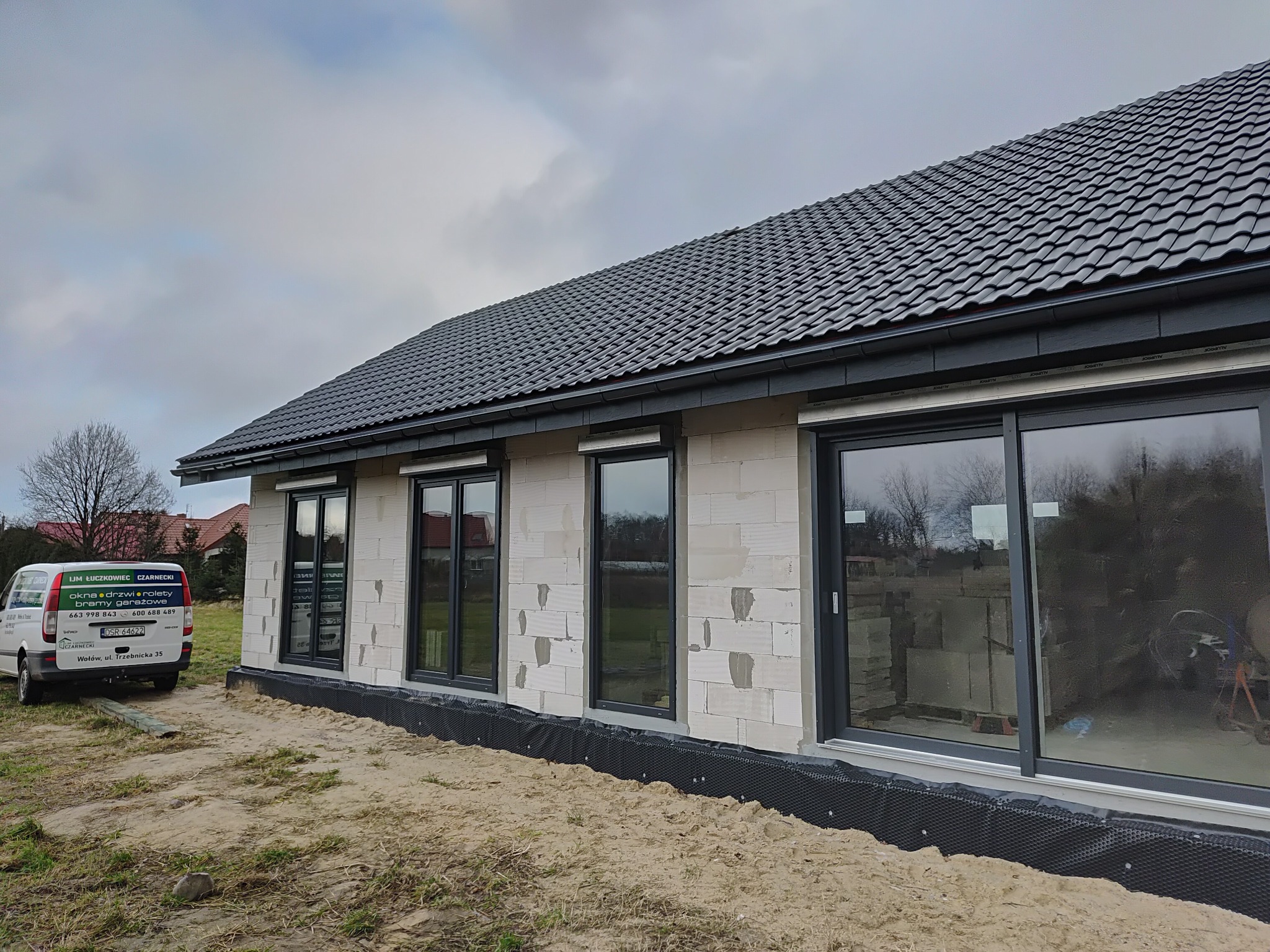 Dom w trakcie budowy z zamontowanymi oknami aluminiowymi w ciemnych ramach. Widoczny samochód dostawczy firmy montującej okna. Dach pokryty ciemną dachówką.