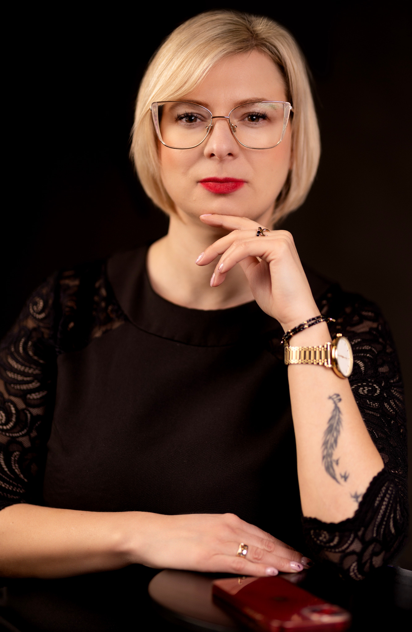 Portret kobiety w okularach, z blond włosami i czerwoną szminką, w czarnej bluzce z koronkowymi rękawami. Na ręce złoty zegarek i tatuaż pióra.