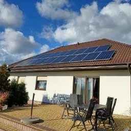 Dom jednorodzinny z panelami słonecznymi na brązowym dachu, widok z tarasu z meblami ogrodowymi w słoneczny dzień z chmurami.