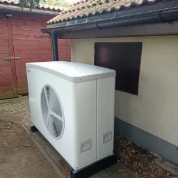 Biała pompa ciepła Daikin zainstalowana przy budynku z brązową stolarką okienną i drewnianą szopą w tle.