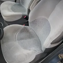 Tapicerka fotela samochodowego z wyraźnymi śladami użytkowania i niewielkimi uszkodzeniami materiału, widoczna kierownica i fragment deski rozdzielczej.
