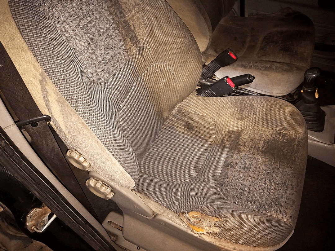 Bardzo zabrudzone i podarte szare siedzenie samochodowe z widocznymi plamami, dziurą w tapicerce, pasem bezpieczeństwa i zagłówkiem w tle. Wnętrze pojazdu w słabym oświetleniu.