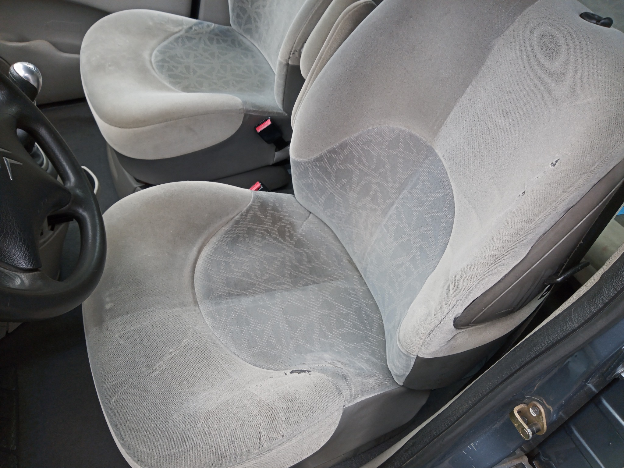 Tapicerka fotela samochodowego z wyraźnymi śladami użytkowania i niewielkimi uszkodzeniami materiału, widoczna kierownica i fragment deski rozdzielczej.