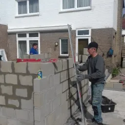 Murarz w czapce i rękawicach, sprawdzający pion ściany z betonowych bloczków za pomocą długiej poziomicy; w tle biały dom z cegły i drugi murarz z poziomicą na murze.