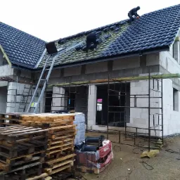 Budowa domu z białego pustaka: dekarze układają ciemną dachówkę na połaci dachu, widoczny ruszt z łat i kontrłat, palety z dachówkami na ziemi.