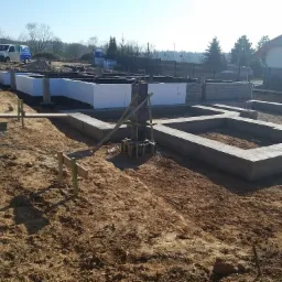 Betonowe fundamenty domu jednorodzinnego w trakcie budowy, z widoczną izolacją termiczną, szalunkami i stertą bloczków betonowych w tle.