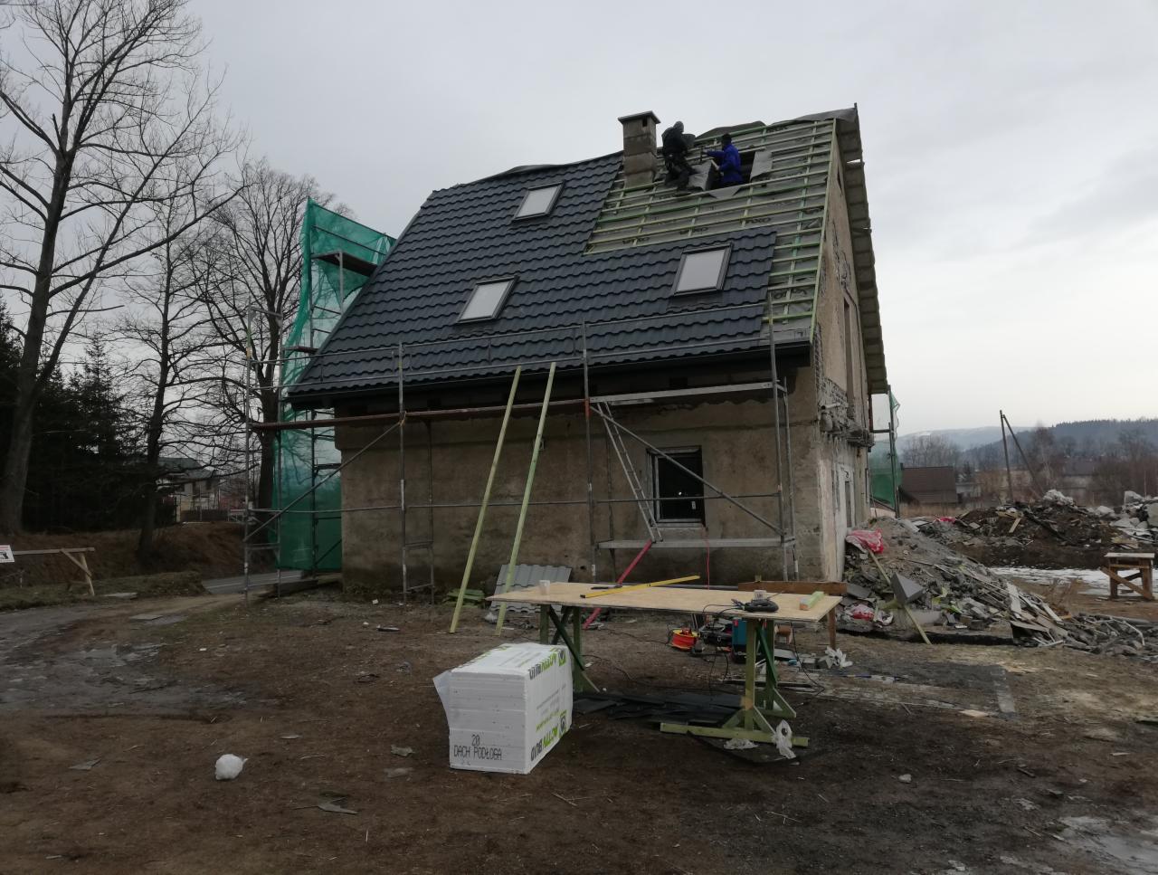 Ekipa dekarzy na rusztowaniu, montująca czarną dachówkę na domu z oknami dachowymi, widoczne narzędzia i materiały budowlane na ziemi.