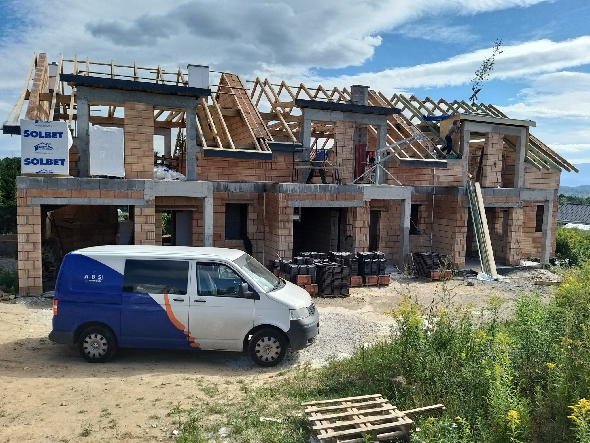 Budowa domu murowanego w stanie surowym otwartym z widoczną konstrukcją dachu, materiały budowlane SOLBET na paletach, oraz samochód dostawczy firmy budowlanej na pierwszym planie.