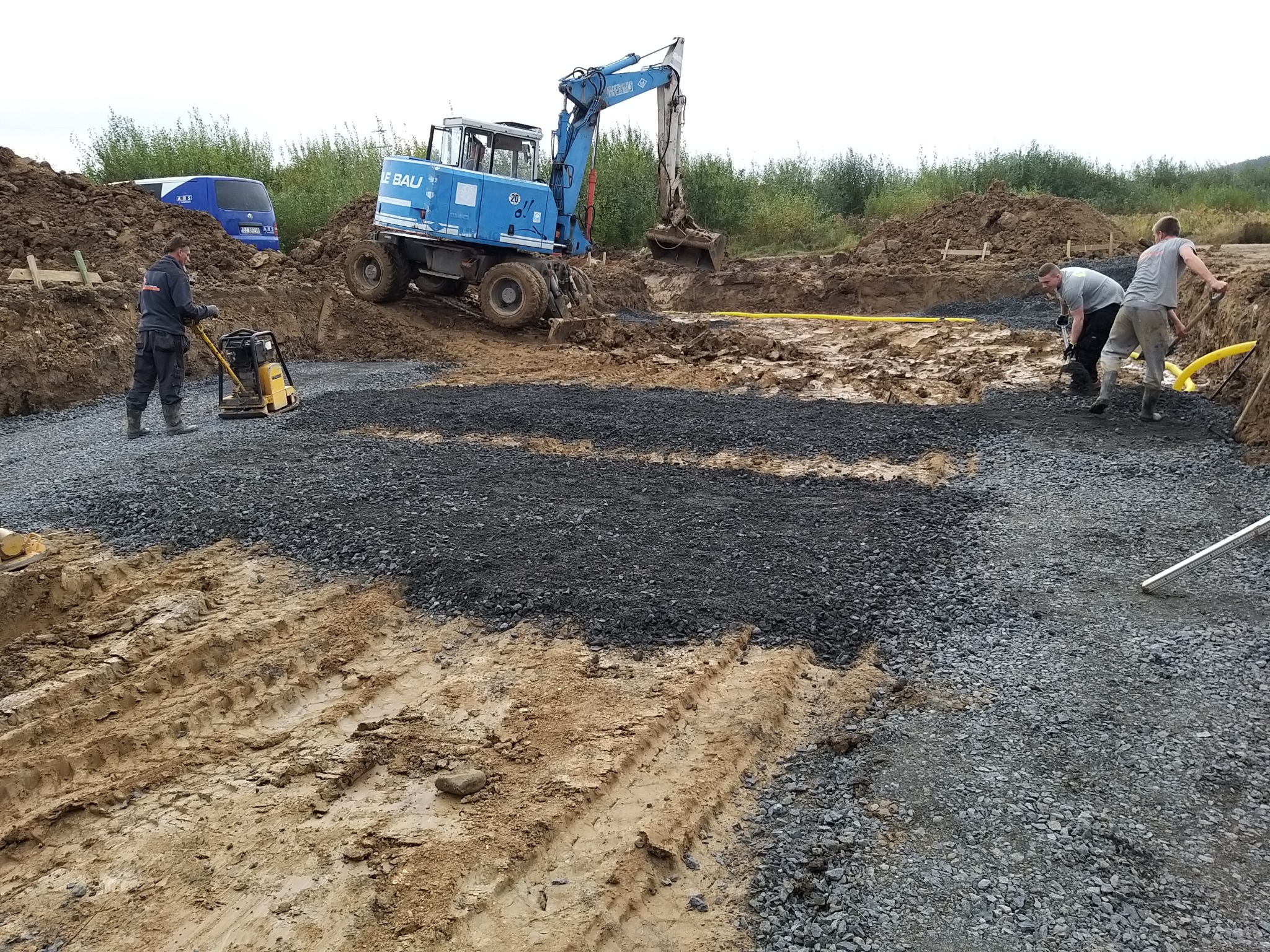 Prace ziemne z użyciem zagęszczarki i koparki na budowie, widoczne wykopane fundamenty i ułożona żółta rura.