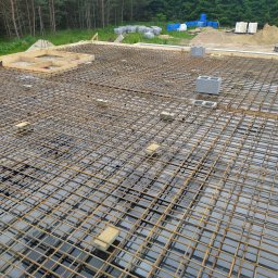 Mak-Bud Paweł Makar - Zbrojenie płyty fundamentowej z siatki stalowej na czarnej hydroizolacji, z drewnianymi szalunkami i betonowymi bloczkami dystansowymi, w tle drzewa i materiały budowlane.