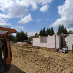 Mak-Bud Paweł Makar - Wznoszenie ścian domu z białych bloczków na fundamencie, widoczne drewniane nadproża okienne, na pierwszym planie otwarty bagażnik pomarańczowego samochodu dostawczego, w tle drzewa iglaste...