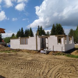 Mak-Bud Paweł Makar - Surowy stan domu w trakcie budowy: ściany z białych bloczków z widocznymi otworami okiennymi i nadprożami, teren wokół z piaskiem i śladami opon, w tle zielone drzewa i błękitne niebo z chmurami.