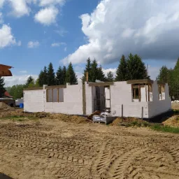 Surowy stan domu w trakcie budowy: ściany z białych bloczków z widocznymi otworami okiennymi i nadprożami, teren wokół z piaskiem i śladami opon, w tle zielone drzewa i błękitne niebo z chmurami.