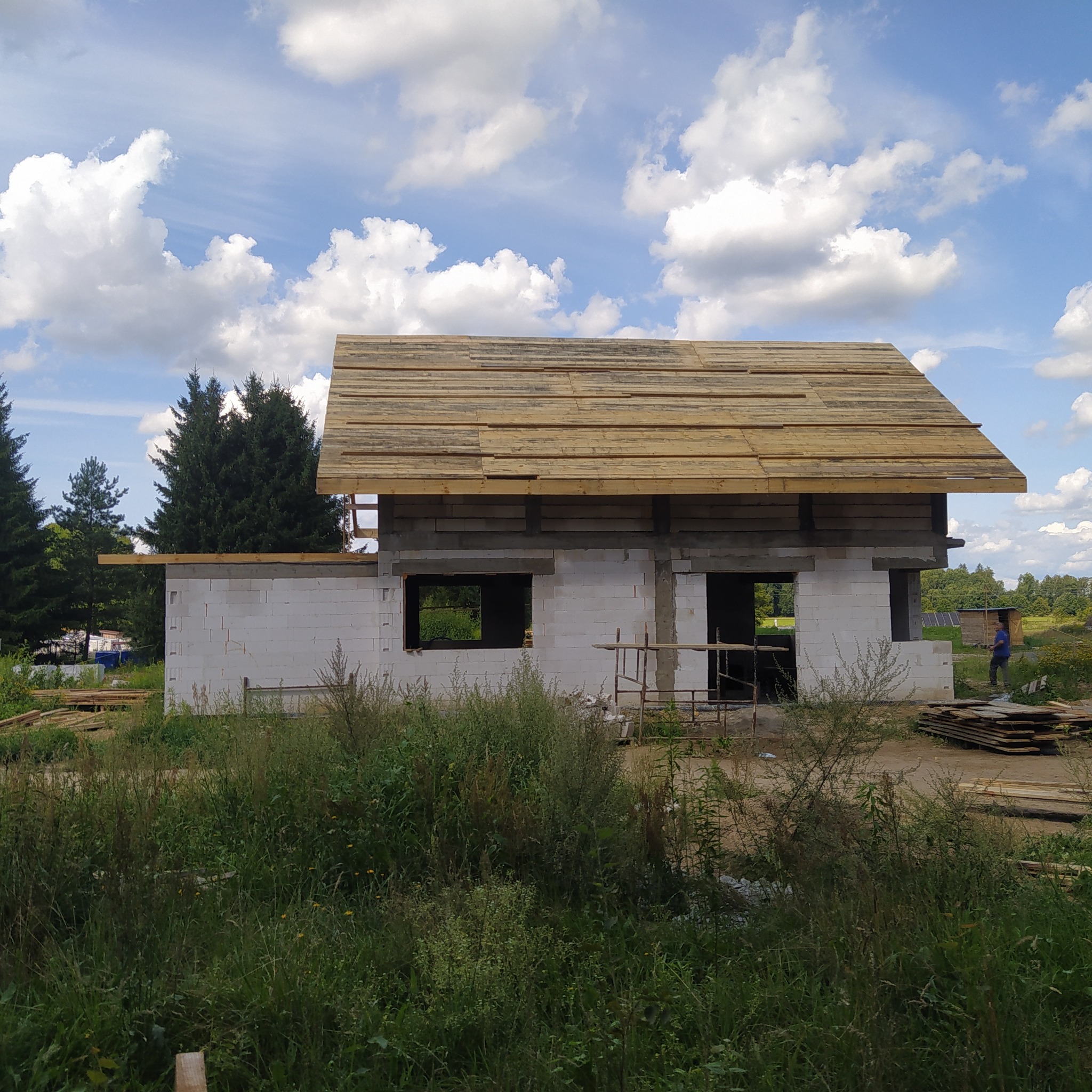 Surowy stan domu w trakcie budowy z częściowo wykonanym drewnianym szkieletem dachu, widoczne białe bloczki ścian, zarośnięte otoczenie i błękitne niebo z obłokami.