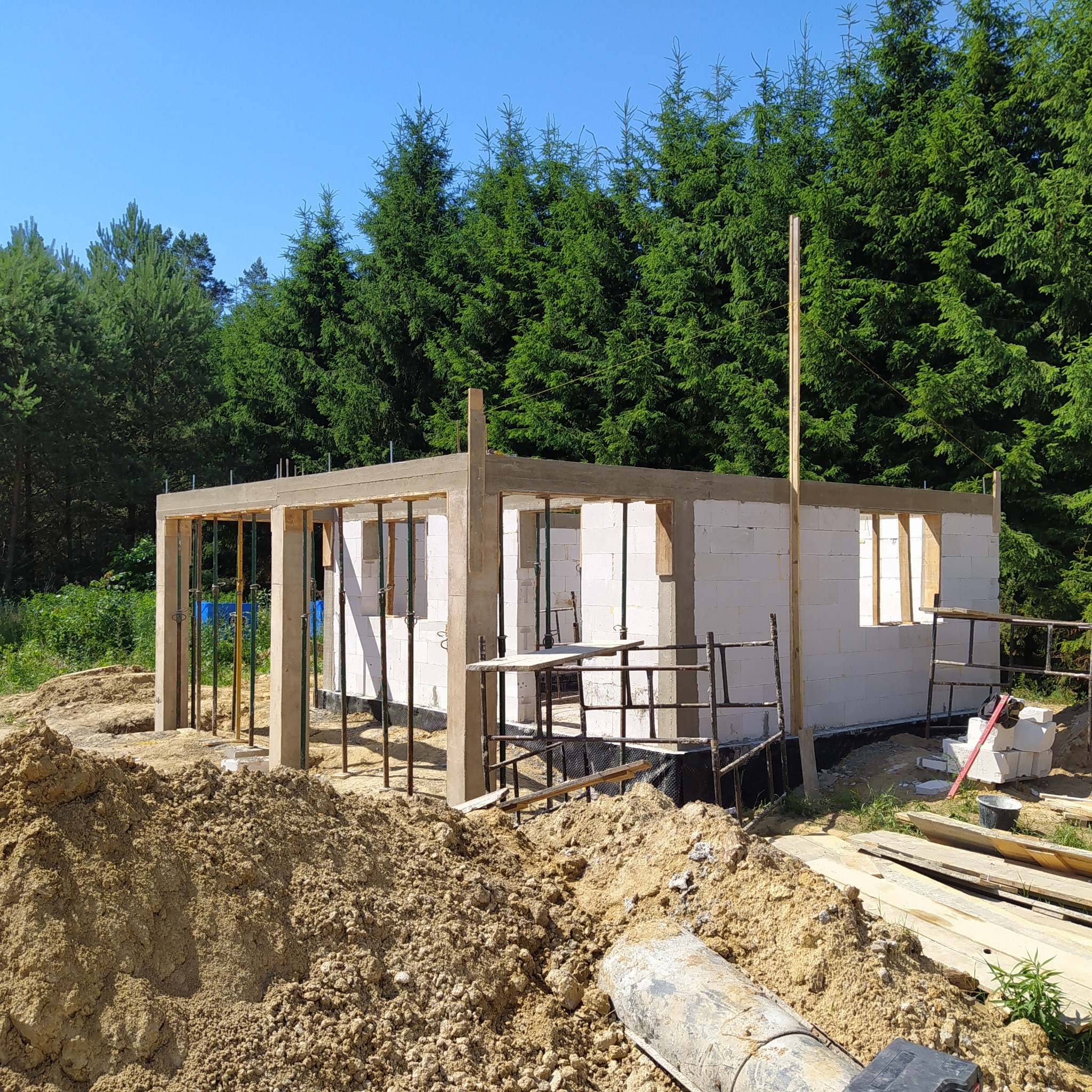 Wznoszenie ścian parterowego domu z betonu komórkowego na tle lasu, widoczne podpory szalunkowe, hałda piasku i narzędzia budowlane.