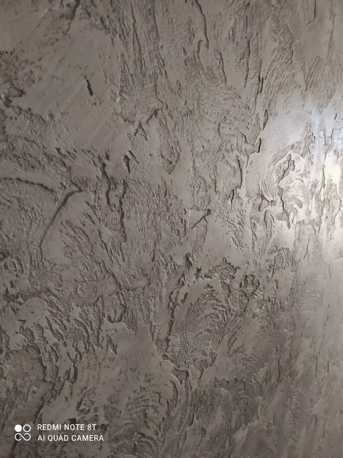 Szpachlowana ściana z wyraźną, nieregularną fakturą w odcieniach szarości, wykonana techniką dekoracyjną.