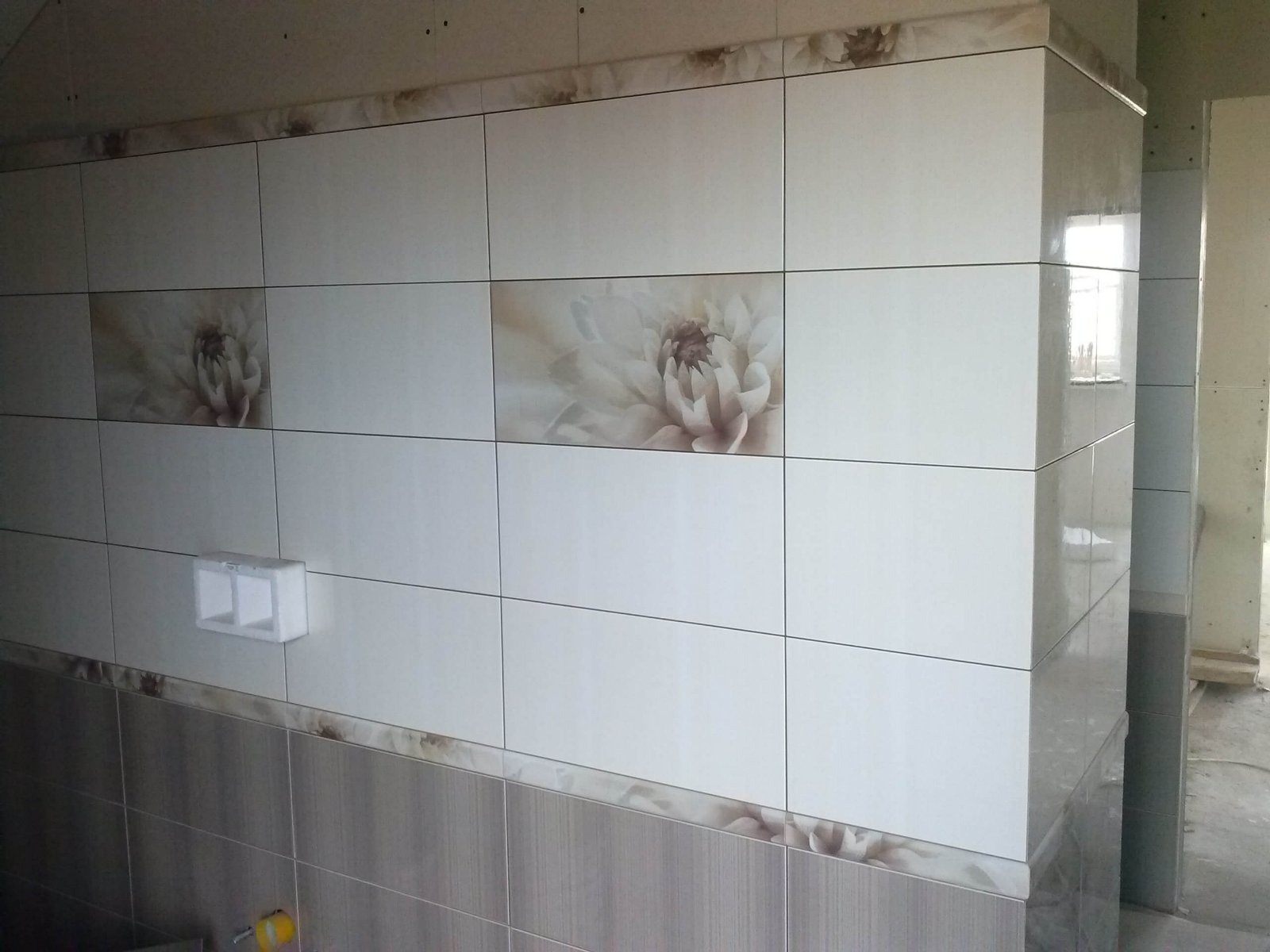 Wykończona ściana w łazience z białymi i szarymi płytkami z motywem kwiatowym, widoczny narożnik i fragment surowego stanu pomieszczenia.