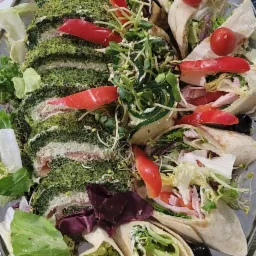 Dekoracyjnie ułożone na srebrnej tacy roladki szpinakowe z nadzieniem i tortille z szynką, warzywami i pomidorami koktajlowymi, ozdobione kiełkami i papryką na stole z motywem liści.