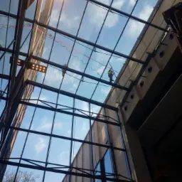 Dwóch pracowników zabezpieczonych linami, wykonujących prace na wysokościach na szklanym dachu budynku z widocznymi elementami konstrukcyjnymi i niebem w tle.