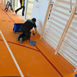 Kobieta w czarnym ubraniu na kolanach, w ż&oacute;łtych rękawicach, czyści białą ścianę w sali gimnastycznej obok drabinek gimnastycznych. Na pomarańczowej podłodze leży niebieska ścierka, a obok stoi...