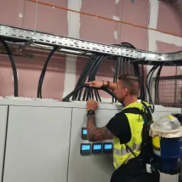 Elektryk w kamizelce odblaskowej układa kable w korycie kablowym nad szafą sterowniczą, widoczna tabliczka ostrzegawcza o ryzyku porażenia prądem.