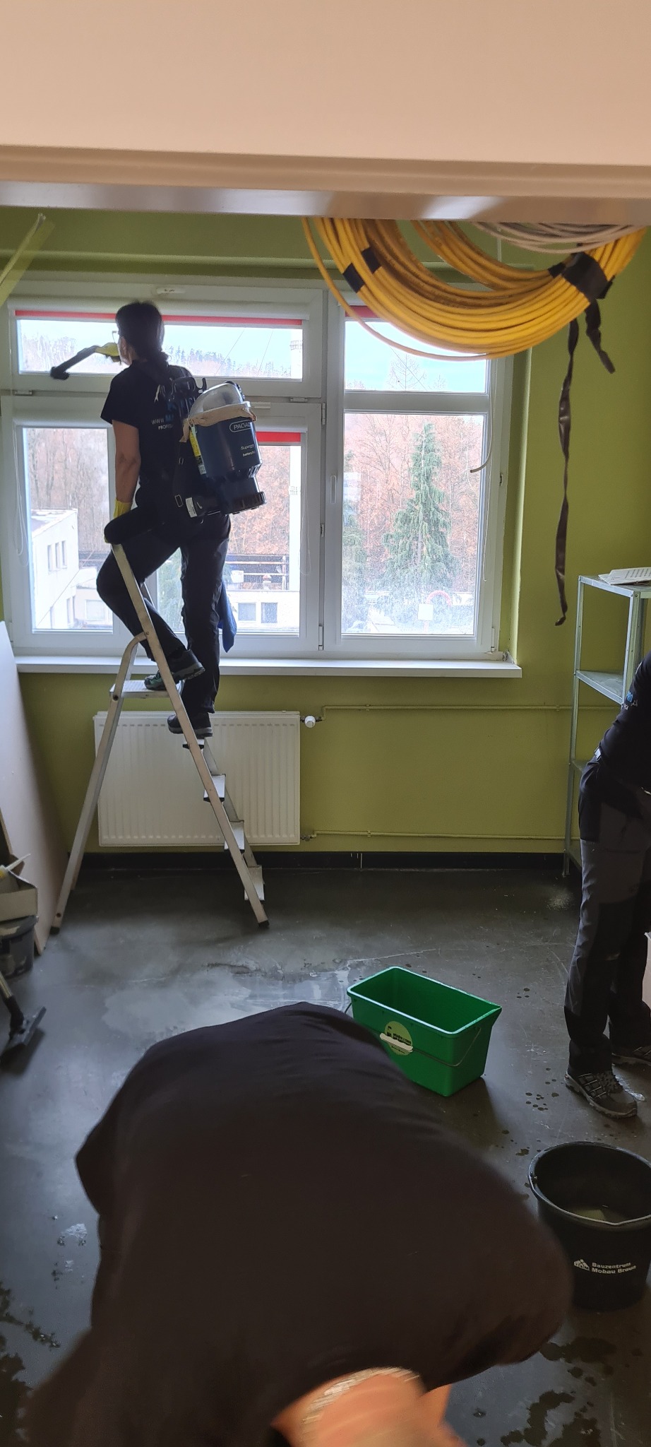 Pracownik na drabinie myje okno od wewnątrz, używając profesjonalnego sprzętu z plecakiem, widoczne wiadro i mokra podłoga.