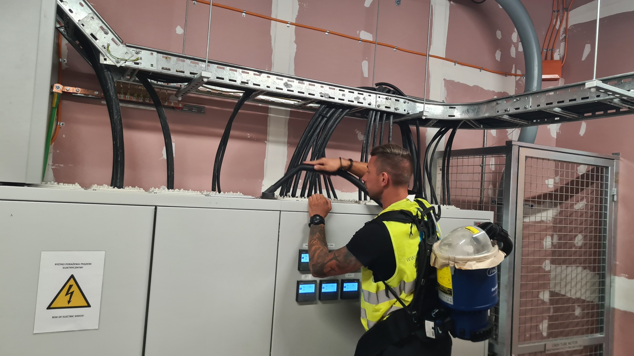 Elektryk w kamizelce odblaskowej układa kable w korycie kablowym nad szafą sterowniczą, widoczna tabliczka ostrzegawcza o ryzyku porażenia prądem.