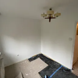 Pokój po malowaniu na biało, zabezpieczony folią malarską i kartonem, widoczny grzejnik i staromodny żyrandol.