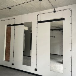 Łukasz Berliński - Wnętrze w trakcie wykańczania: białe ściany, szara podłoga, trzy lustra z czarnymi oprawami zawieszone na ścianach, widoczne kable elektryczne prowadzone po ścianach i suficie, fragment ściany...