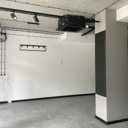 Łukasz Berliński - Wnętrze w Brwinowie w stanie deweloperskim, świeżo po wykonaniu prac wykończeniowych: białe ściany, szara podłoga z płytek, widoczne instalacje elektryczne pod sufitem i fragment ściany z cegły