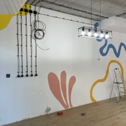 Wnętrze biura w trakcie malowania artystycznego: białe ściany z abstrakcyjnymi, kolorowymi wzorami, drabina, widoczne przewody elektryczne i oświetlenie sufitowe.