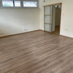 Puste pomieszczenie biurowe z nową podłogą z paneli w odcieniach drewna, jasnymi ścianami i przesuwanymi drzwiami z matowego szkła.